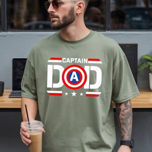 K&ouml;nnte beinhalten: Olivgr&uuml;nes T-Shirt mit dem Text "CAPTAIN DAD" in wei&szlig;en Blockbuchstaben. Das "D" in "DAD" hat einen blauen Kreis mit einem wei&szlig;en "A" darin und rot-wei&szlig;e Streifen. Darunter drei wei&szlig;e Sterne.