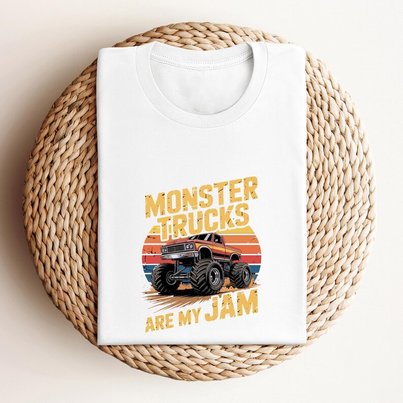 May include: White t-shirt featuring a graphic of a red and brown monster truck with large black tires, set against a sunset design. The text "MONSTER TRUCKS ARE MY JAM" is printed above and below the truck in a distressed yellow font.