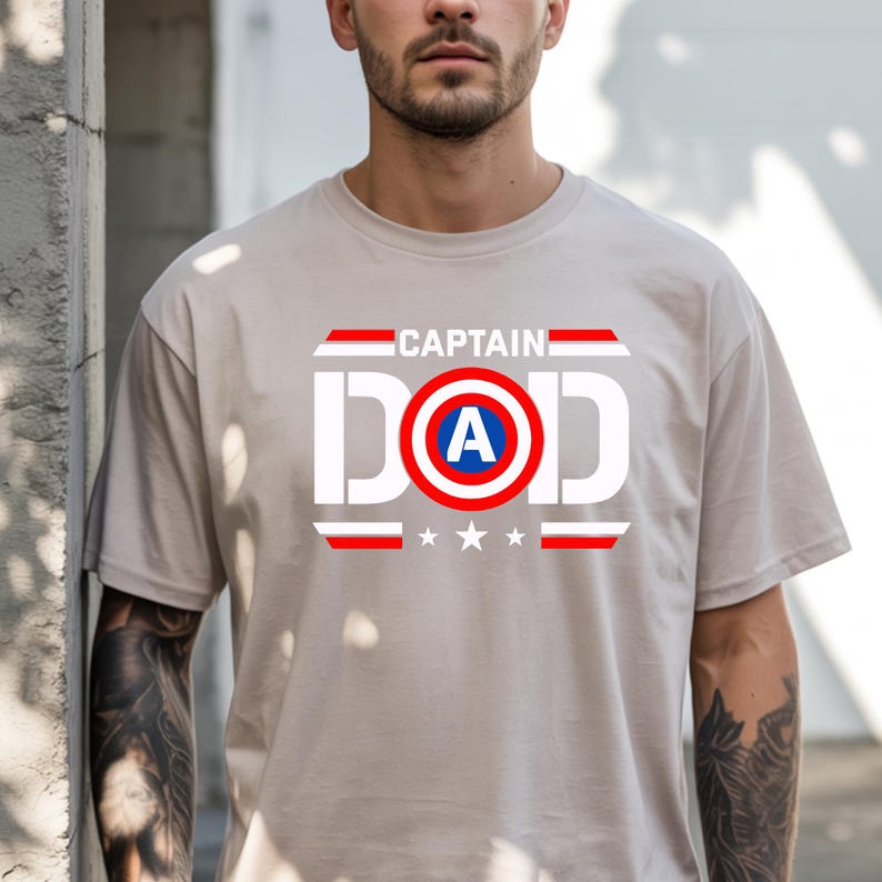 K&ouml;nnte beinhalten: Hellbeige T-Shirt mit dem wei&szlig;en Schriftzug "CAPTAIN DAD". Das "A" in "DAD" befindet sich in einem rot-wei&szlig;-blauen Schild. Darunter befinden sich drei wei&szlig;e Sterne und rote Streifen, ein Superhelden-Design.