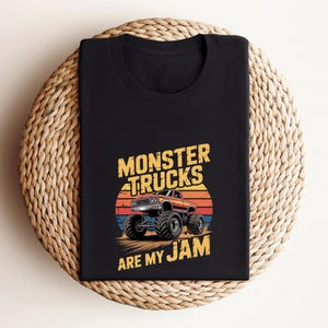 May include: Black t-shirt featuring a graphic of a monster truck with large tires, set against a sunset backdrop. The text "MONSTER TRUCKS ARE MY JAM" is printed above and below the truck in yellow. The shirt is folded on a woven surface.