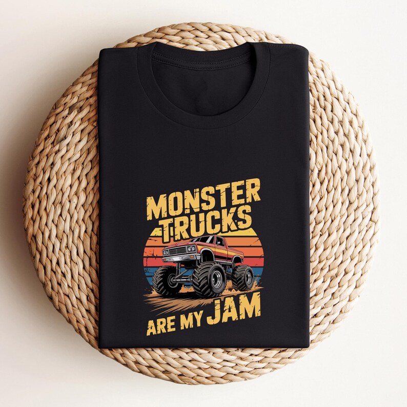 May include: Black t-shirt featuring a graphic of a monster truck with large tires, set against a sunset backdrop. The text "MONSTER TRUCKS ARE MY JAM" is printed above and below the truck in yellow. The shirt is folded on a woven surface.