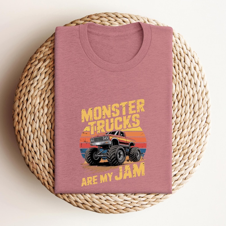 May include: A dusty rose t-shirt featuring a graphic of a monster truck with the text "MONSTER TRUCKS ARE MY JAM". The design includes a sunset backdrop with orange, red, and blue stripes. The shirt is folded on a woven, circular placemat.