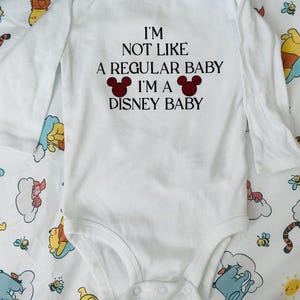 Könnte beinhalten: Weißer langärmliger Baby-Strampler mit schwarzem Text, der "Ich bin nicht wie ein normales Baby Ich bin ein Disney-Baby" lautet, mit zwei roten Mickey-Mouse-Ohren auf beiden Seiten des Textes.
