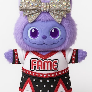 May include: A plush purple creature with a large jeweled bow on its head. It wears a cheerleader outfit with the word "FAME" on the front. The outfit is white, black, and red.