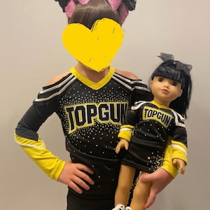 May include: A young girl wearing a black and yellow cheerleading uniform with the word "TOP GUN" on the front, holding a doll wearing a matching cheerleading uniform. Both are wearing black skirts and black sneakers.