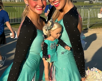 Könnte beinhalten: Zwei lächelnde Cheerleaderinnen in türkis-schwarzen Uniformen mit einer Puppe in passendem Outfit. Die Uniformen haben schwarze Ärmel mit weißen Punkten und sind mit Pailletten verziert. Sie halten Pompons.