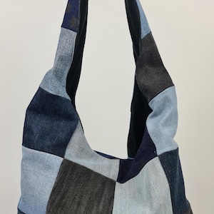 May include: A patchwork denim shoulder bag in various shades of blue and grey. The bag has a large, rounded shape with a wide strap and is made from squares of recycled denim. The interior and strap are black.