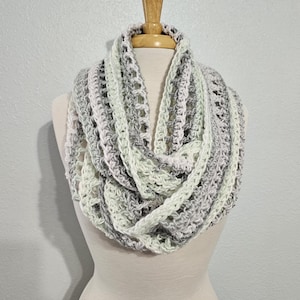 May include: A chunky crochet infinity scarf featuring a variegated yarn pattern in shades of cream, light gray, and soft white. The accessory is crafted with an open, airy stitch design that creates a textured, layered appearance around the neck.