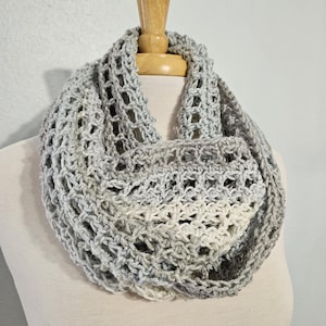 May include: A light gray and white infinity scarf featuring an open, airy crochet stitch pattern. The yarn transitions between shades of gray and off-white, creating a textured, looped design that drapes in a circular, continuous form.