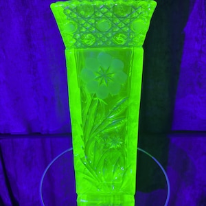 May include: A tall, square-shaped fluorescent yellow glass vase glowing under ultraviolet light. The top rim features a textured cross-hatch and circle pattern, while the body displays an etched floral design with a central star-shaped flower.