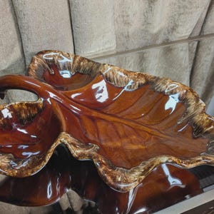 May include: A large, glossy, leaf-shaped serving dish in shades of brown and tan. The ceramic dish has a curved handle and a textured rim, reflecting light. This decorative piece is suitable for serving snacks or as a decorative accent.