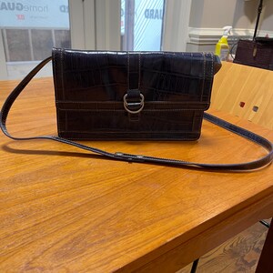 May include: A dark navy blue crossbody bag with a crocodile skin texture. The bag has a rectangular shape with a flap closure, a silver-colored D-ring, and a long, thin strap. The bag is sitting on a wooden table.