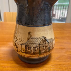May include: A ceramic pitcher with a dark blue rim and handle, and a tan body. The body features a hand-painted cabin scene with a tree. The pitcher is sitting on a wooden table.