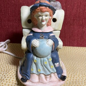May include: A ceramic witch figurine seated in a chair, holding a blue crystal ball. The witch wears a blue hat with an orange band, a blue dress, and a light blue skirt with yellow stars. A gray cat sits behind her. The figurine has a power cord.