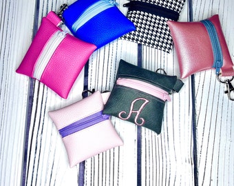 May include: Six small zippered pouches in various colors and patterns. The pouches are made of leather and fabric and have a zipper closure. The pouches are arranged on a white wooden surface.