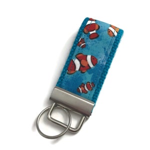 May include: A turquoise blue key chain with a silver key ring and a fabric strap featuring a repeating pattern of orange and white clownfish on a blue background.