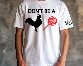 Może przedstawiać: Biała koszulka z nadrukiem "DON'T BE A" w kolorze czarnym. Po lewej stronie koszulki znajduje się czarna sylwetka koguta, a po prawej stronie czerwony lizak. Na prawym rękawie nadrukowany jest czarny napis "US".
