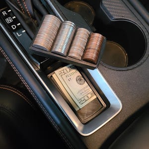 May include: A black leather car console with a coin holder and a twenty pound note in a leather wallet. The coin holder has four compartments, each filled with a stack of coins. The coins are arranged in order of size, with the largest coins at the back and the smallest coins at the front.