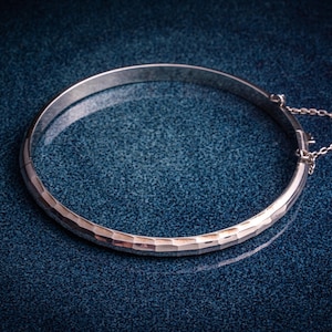 May include: A silver hinged bangle bracelet with a textured surface. The bracelet has a simple design and a small chain for added security. The bangle is displayed on a dark blue textured surface.
