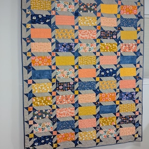 May include: A large, colorful quilt with a patchwork design. The quilt features rectangular blocks in shades of yellow, peach, navy, and floral patterns, set against a gray background. Dark blue star shapes frame the edges. The quilt is hanging on a wall.