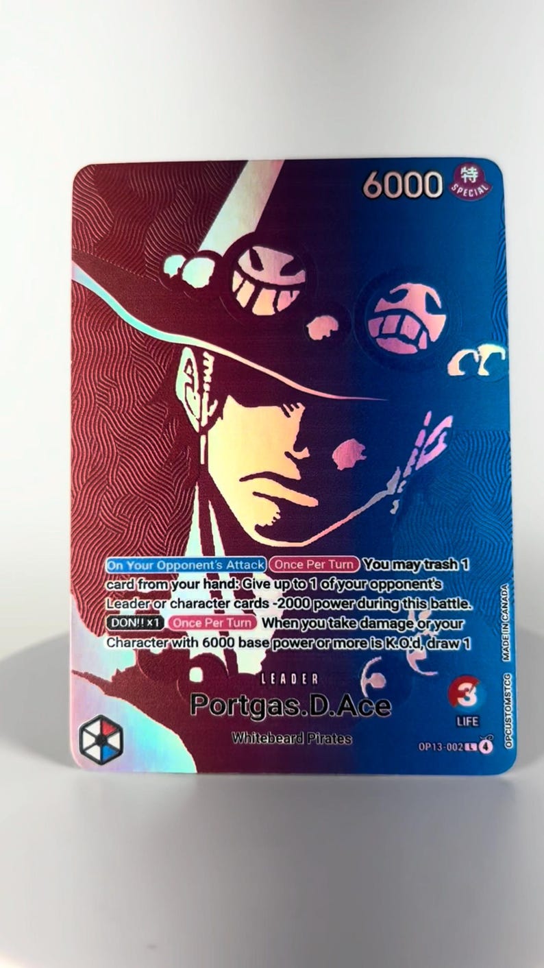 May include: A holographic trading card featuring Portgas D. Ace from the Whitebeard Pirates. The card has a blue and red color scheme with the character's image and game text. The card has a power level of 6000 and a life value of 3.