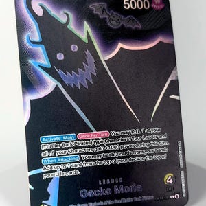 Puede incluir: Una carta de líder de Gecko Moria del juego de cartas One Piece. La carta presenta una silueta oscura de un personaje con un murciélago y el número 5000. El texto incluye instrucciones del juego y el nombre de la carta.