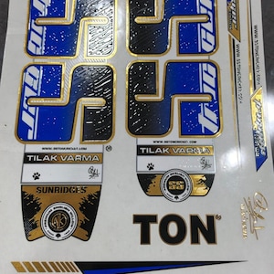 May include: Cricket bat stickers with blue, black, and gold accents. The stickers feature the text "SS" and "TON", along with the name "TILAK VARMA" and the word "SUNRIDGES". The stickers are on a white background.