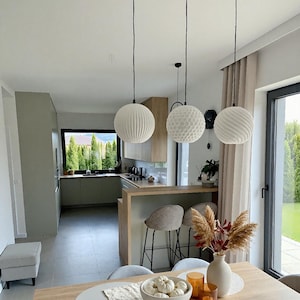 May include: A contemporary kitchen and dining space with three white spherical pendant lights. The lights are suspended above a wooden dining table with a bowl of white pumpkins and decorative items. A kitchen island with bar stools is visible.