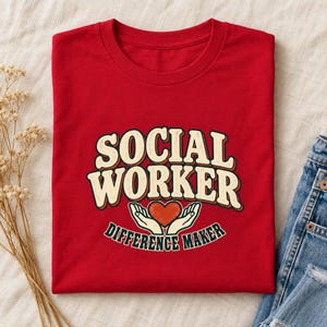 Könnte beinhalten: Ein rotes Kurzarm-T-Shirt mit dem Text SOCIAL WORKER in geschwungener, cremefarbener Schrift mit dunkler Umrandung. Unter dem Text befindet sich eine Illustration von zwei Händen, die ein rotes Herz halten, mit dem Schriftzug DIFFERENCE MAKER.
