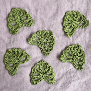 Peut inclure: Six feuilles de Monstera en crochet vert faites à la main sont disposées sur un tissu violet clair. Les feuilles présentent un motif ajouré détaillé, mettant en valeur le travail de crochet. Les feuilles sont d'un vert vif.