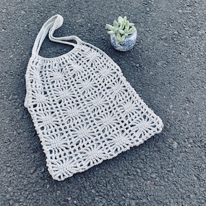 May include: A gray crocheted tote bag with a circular pattern. The bag is lying on a gray asphalt surface.