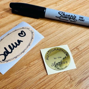 May include: A black Sharpie fine point marker rests on a wooden surface next to two stickers. One sticker is oval-shaped with the name "Selma" and a heart. The other is a gold circle that says "Autographed by the Author."