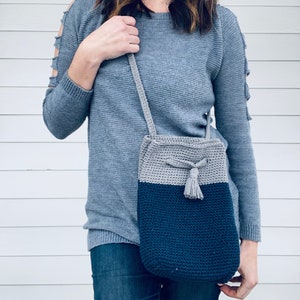 May include: A gray and blue crocheted crossbody bag with a tassel. The bag is worn by a person wearing a gray sweater.