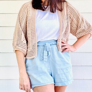 May include: A beige, open-front crochet cardigan with short sleeves. The cardigan is worn over a white t-shirt and paired with light blue shorts. The crochet design features a loose, open weave.