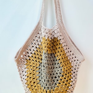 Könnte beinhalten: Eine gehäkelte Tragetasche in Beige und Gelb mit langen Trägern. Die Tasche hat ein kreisförmiges Muster in Beige-, Gelb- und Grautönen. Das offene Webdesign ist über die gesamte Tasche sichtbar und erzeugt ein strukturiertes Aussehen. Die Tasche hängt vor einem weißen Hintergrund.