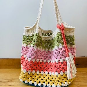 May include: A crocheted tote bag with a colorful striped design. The bag has a green, pink, orange, and yellow pattern. The bag has a white crocheted handle and a white tassel.