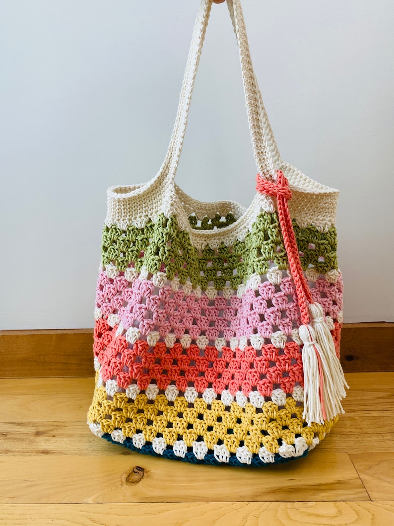 May include: A crocheted tote bag with a colorful striped design. The bag has a green, pink, orange, and yellow pattern. The bag has a white crocheted handle and a white tassel.
