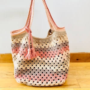 May include: A crocheted tote bag with pink, beige, and brown stripes. The bag has a long strap and a tassel.