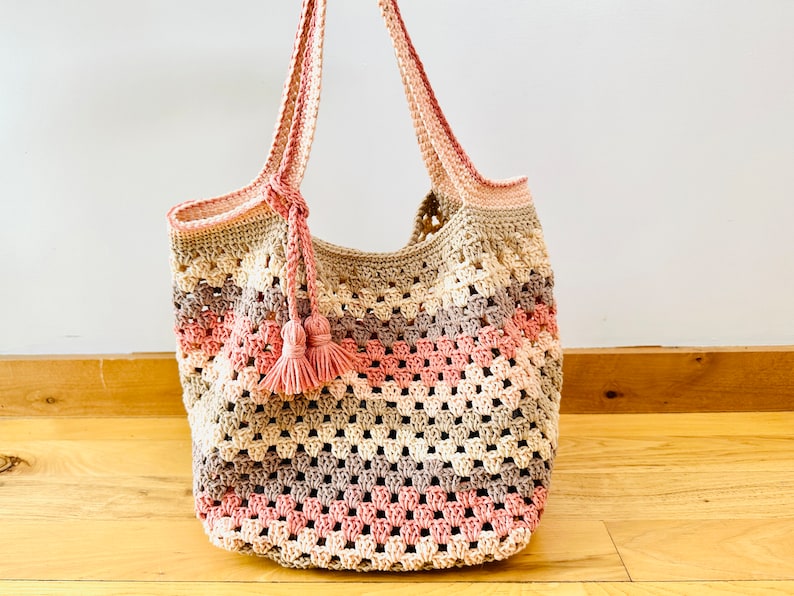 May include: A crocheted tote bag with pink, beige, and brown stripes. The bag has a long strap and a tassel.
