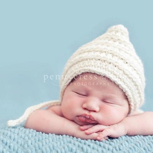 May include: A newborn baby wearing a cream-colored knitted hat with ear flaps, resting on a light blue blanket. The baby is sleeping with its eyes closed, arms crossed, and lips slightly puckered. The background is a soft blue.