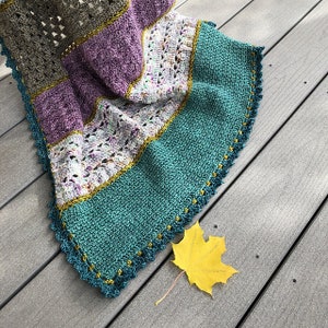 Könnte beinhalten: Eine gehäkelte Decke mit einem mehrfarbigen Streifenmuster in Braun-, Lila-, Weiß-, Grün- und Goldtönen. Die Decke hat einen gewellten Rand mit einem goldenen Besatz. Ein gelbes Blatt liegt auf der Holzoberfläche neben der Decke.