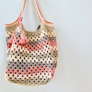 May include: A crocheted tote bag with a pink, beige, and brown color scheme. The bag has a wide, open top and two long straps with tassels.