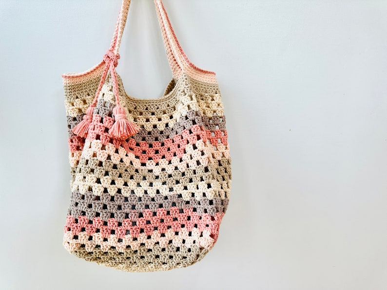 May include: A crocheted tote bag with a pink, beige, and brown color scheme. The bag has a wide, open top and two long straps with tassels.