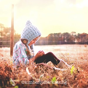 Puede incluir: Un niño con una capucha de ganchillo azul claro con un detalle de botón, sentado en un campo de hierba seca. El niño lleva una camisa estampada y leggings oscuros. La imagen está bañada por la cálida luz del sol.