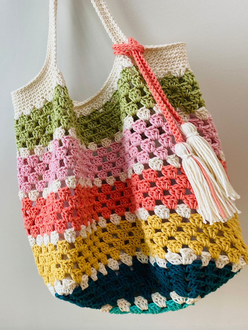 May include: A colorful crocheted tote bag with a white tassel. The bag is made with different colored yarn in stripes of green, pink, orange, yellow, and blue.