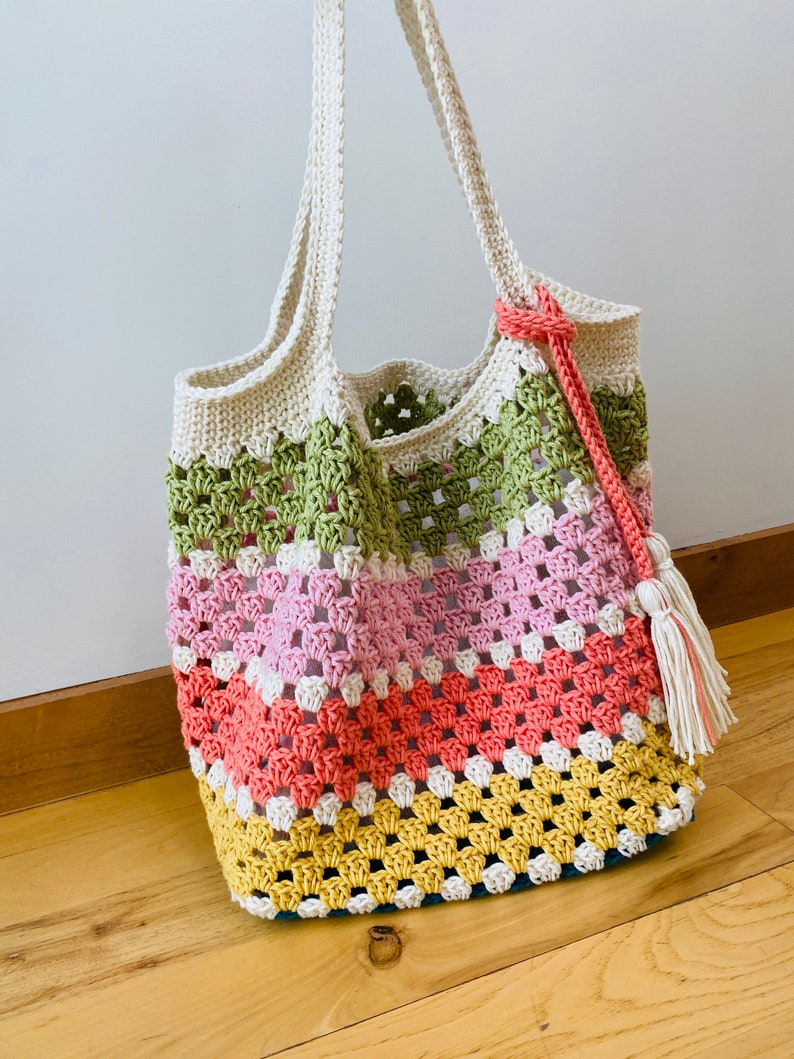 May include: A crocheted tote bag with a striped pattern in green, pink, orange, and yellow. The bag has a white handle and a white tassel.