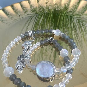 Op de afbeelding: Drie armbanden met kralen, een zilveren bedel en een grote ovale maansteenring. De armbanden hebben heldere en grijze kralen, met zilveren accenten. De sieraden worden gepresenteerd op een goudkleurige decoratieve schaal.