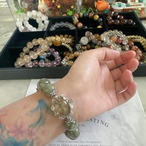 Könnte beinhalten: Ein Armband mit großen, runden, grünen Perlen und einem zentralen, oval geformten Stein. Das Armband befindet sich an einem Handgelenk mit einem floralen Tattoo. Weitere Armbänder sind in einer schwarzen Schale zu sehen. Der Text auf dem Papier lautet: „Es gibt keine Zeit wie die Gegenwart.“
