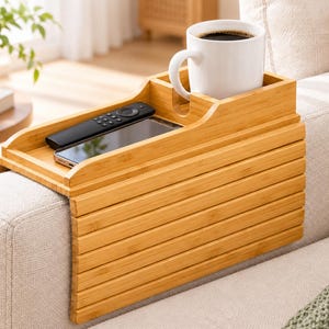 May include: A light brown bamboo sofa arm tray with a flexible, slatted design that drapes over the armrest. It features a built-in cup holder containing a white mug, a rectangular slot for a remote control, and a flat surface holding a smartphone.
