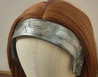 May include: A wig with short, auburn hair and a silver-colored, metallic headband. The wig is displayed on a mannequin head. The headband has a geometric design and appears to be made of a lightweight material.
